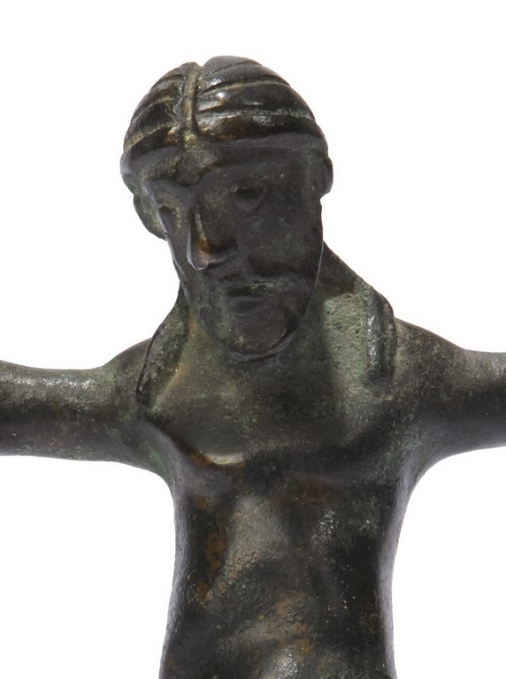 Christ en bronze patiné. Le dos creux, il est représenté vivant, les yeux en relief, les cheveux séparés par une raie médiane retombant sur les épaules, les bras écartés terminés par de larges mains, les jambes parallèles presque entièrement recouvertes par un important perizonium aux amples plis.